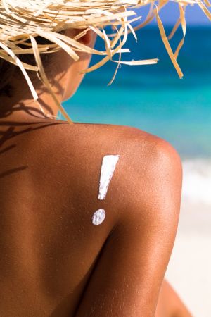 Auf der gebräunten Schulter einer Frau ist ein Ausrufezeichen aus Sonnencreme, sie trägt einen Strohhut.