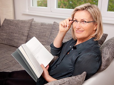 Frau sitzt mit einem Buch auf dem Sofa und lächelt in die Kamera.