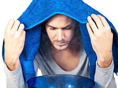 Mann hat ein blaues Handtuch über dem Kopf und beugt sich über eine Schüssel mit dampfendem Wasser.