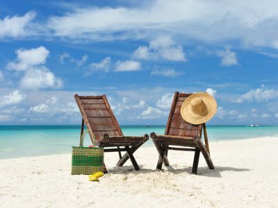 Weißer Sand, türkisfarbenes Wasser, hellblauer Wolkenhimmel, zwei Strandstühle mit Hut und Tasche