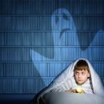 Ein Junge sitzt mit seiner Taschenlampe unter der Bettdecke, in seinem Rücken ist ein Gespenst zu sehen.