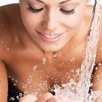 Lächelnde junge Frau wäscht ihr Gesicht mit Wasser.