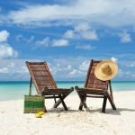 Weißer Sand, türkisfarbenes Wasser, hellblauer Wolkenhimmel, zwei Strandstühle mit Hut und Tasche