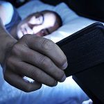 Ein junger Mann liegt im Bett und wird vom blauen Licht seines Smartphones beleuchtet.